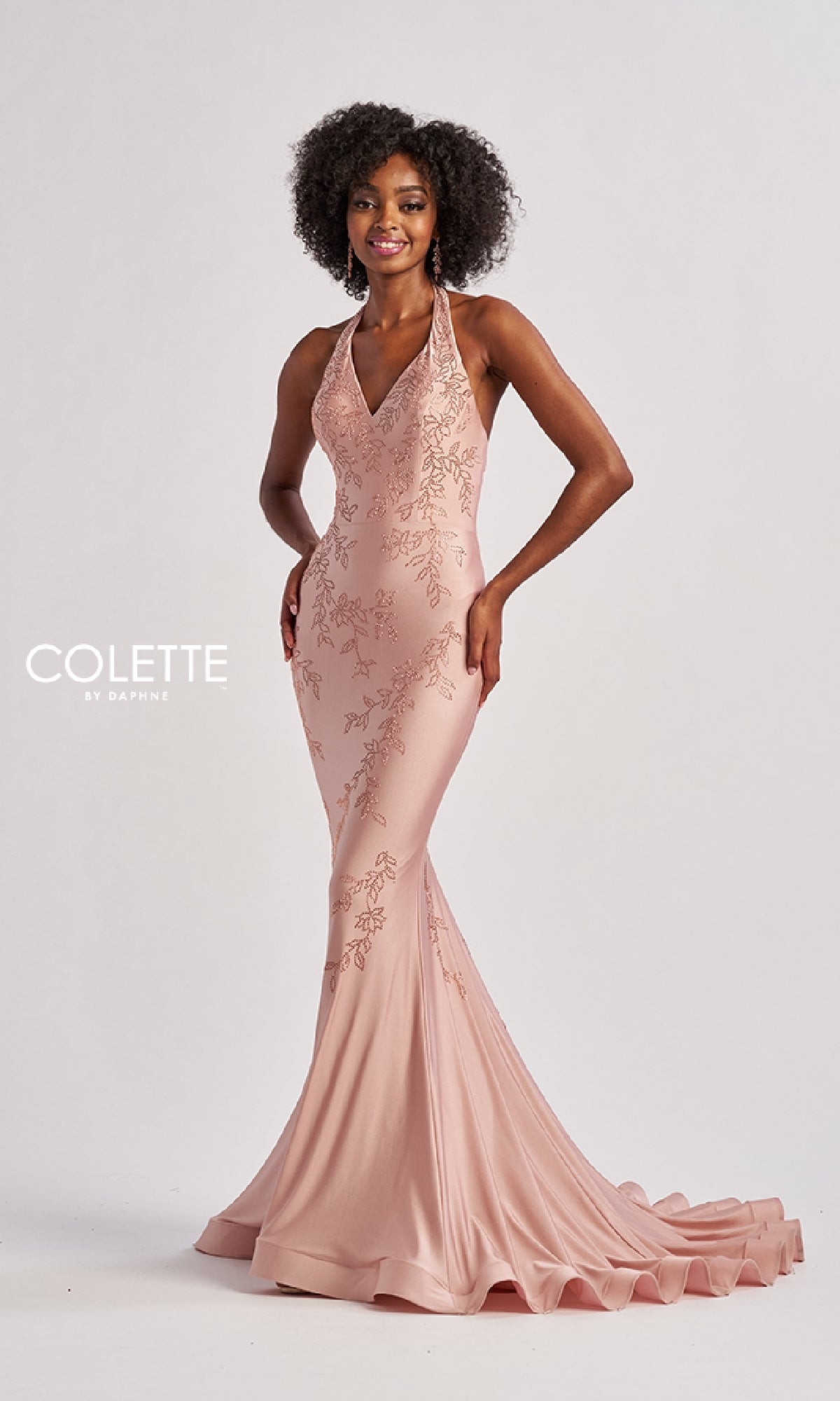 A woman stands against a plain backdrop wearing the Colette CL8580 long prom dress—a pink, halter v-neck, floor-length mermaid gown with floral embroidery, beaded sequin appliques, and a small train.